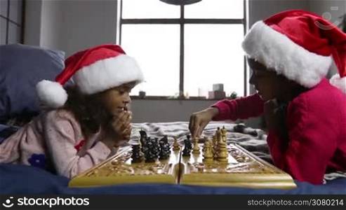 Cute small girls in santa hats playing chess lying on the bed and thinking intensely during christmas time. Sweet mixed race sisters spending leisure together at home playing board game on Xmas eve. Dolly shot. Slow motion.