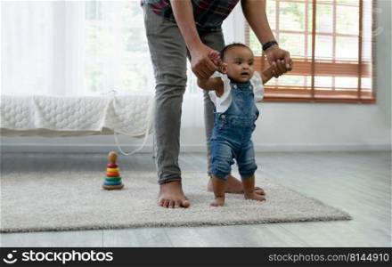 Cute smiling African baby girl learning to walk with father support and holding her hands. Little toddler child taking first steps with parents help at bedroom at home with love and care