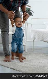 Cute smiling African baby girl learning to walk with father support and holding her hands. Happy little toddler child taking first steps with parents help at bedroom at home with love and care