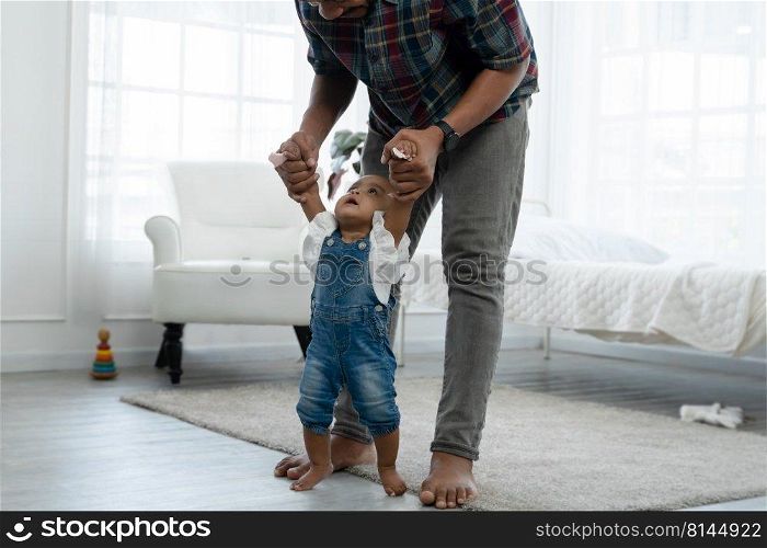 Cute smiling African baby toddler girl looking at father face while learning to walk and taking first steps with help of dad and holding her hands with love and care at bedroom at home