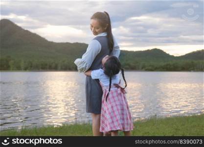 Cute smiling little girl hugging her mother from behind having fun together by the lake. Happy family, mother and little daughter playing feeling happy in the park.