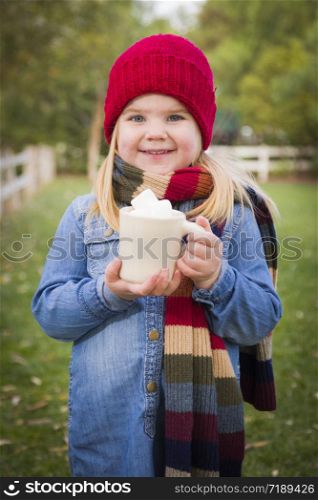 Cute Smiling Young Girl Wearing Hat and Scarf Holding Cocoa Mug with Marsh Mallows Outside.