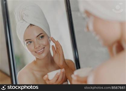 Cute smiling young woman applies beauty cream on face, smiles pleasantly, has minimal makeup, wears wrapped towel on head, looks in mirror, poses in bathroom. Wellness, beauty treatments concept