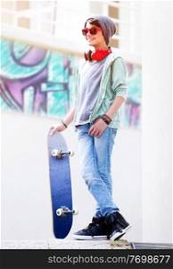 Cute teen boy with skateboard outdoors, standing on the street with different colorful graffiti on the walls, hipster style, cool teen fashion