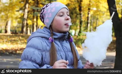 cute teen girl eats tasty candy floss in beautiful autumn city park, closeup