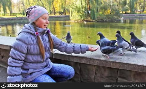 cute teen girl siting feeds pop-corn pigeons from hand in autumn city park near lake, closeup