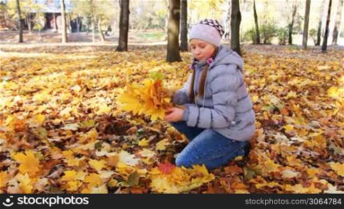 cute teen girl sits on her knee and collects yellow leaves falling from trees in beautiful autumn city park
