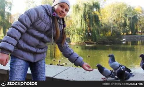 cute teen girl standing feeds pop-corn pigeons from hand in autumn city park near lake