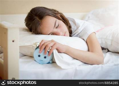 Cute teenage girl holding alarm clock under pillow
