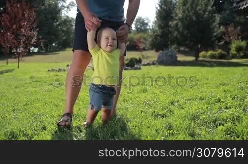 Cute toddler boy with blue eyes learning to walk with father&acute;s help on green grass in summer park. Adorable infant child making his first steps with the help of caring father on grassy lawn on a sunny day. Slow motion. Steadicam stabilized shot.