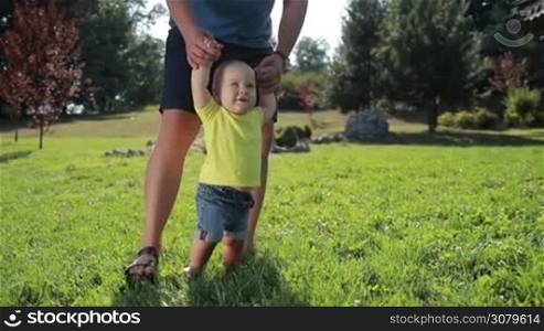 Cute toddler boy with blue eyes learning to walk with father&acute;s help on green grass in summer park. Adorable infant child making his first steps with the help of caring father on grassy lawn on a sunny day. Slow motion. Steadicam stabilized shot.