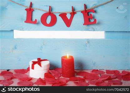 Cute white gift box with tied bow and a red candle, surrounded by soap rose petals and the word love spelled with red paper letters tied to a string.
