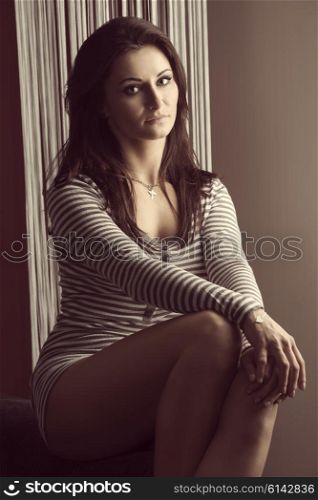 cute woman brunette , looking in camera , with a short dress sitting near a window , she is posing in natural light