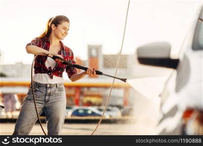 Cute woman cleans car wheels with high pressure water gun. Young lady on self-service automobile wash. Outdoor vehicle washing at summer day. Cute woman cleans car wheels with water gun