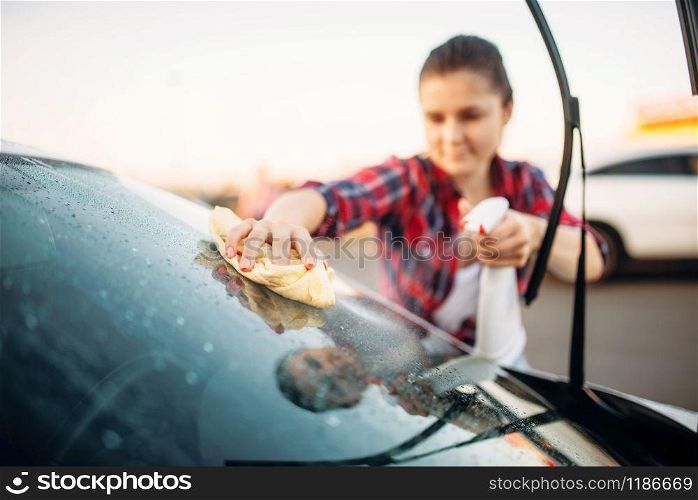 Cute woman cleans front glass of the car with sponge and spray, carwash. Lady on self-service automobile washing. Outdoor vehicle cleaning at summer day