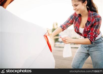 Cute woman cleans rear lights of the car with sponge and spray, carwash. Lady on self-service automobile washing. Outdoor vehicle cleaning at summer day