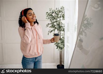Cute woman in headphones listening to music and drinks coffee. Pretty lady in earphones relax in the room, female sound lover resting. Woman listening to music and drinks coffee