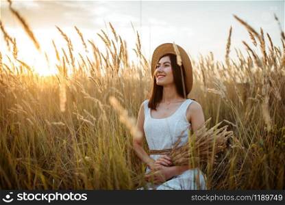 Cute woman in white dress and straw hat holds wheat bouquet in the field. Pretty girl on summer meadow. Woman in dress and straw hat holds wheat bouquet