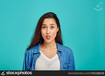 Cute woman shows her tongue, blue background, positive emotion. Face expression, female person looking on camera in studio, emotional concept, feelings. Cute woman shows her tongue, positive emotion