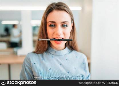 Cute woman with brush in her teeth poses in beauty studio. Cosmetic salon. Professional make up
