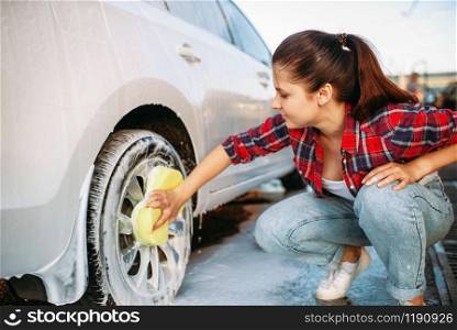 Cute woman with sponge scrubbing vehicle wheel with foam, car wash. Lady on self-service automobile washing. Outdoor carwash at summer day