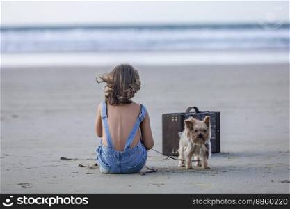 Cute Yorkshire Terrier dog on leash standing near anonymous girl in overall denim shorts and suitcase during summer vacation on Famara Beach in Lanzarote, Spain. Yorkshire Terrier near girl on beach