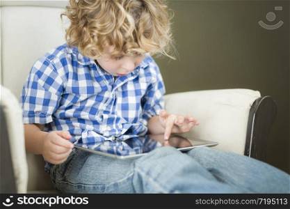 Cute Young Blond Boy Using His Computer Tablet in a Chair.