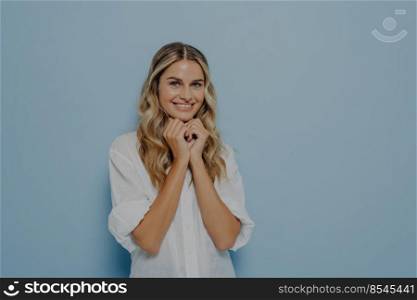Cute young blonde woman with wavy perfect hairstyle folded hands under her chin and waiting for desired gift in excitement, dressed in white shirt. Happy facial expression and positive emotions. Cute young blonde woman with wavy hairstyle folded hands under her chin and waiting for desired gift