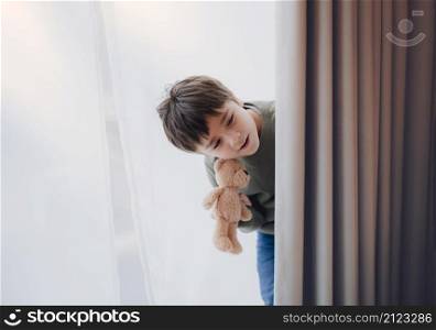 Cute young boy holding teddy bear standing behind lace curtain with bright light in morning, Happy kid with smiling face on window sill and plays at hide-and-seek behind curtain
