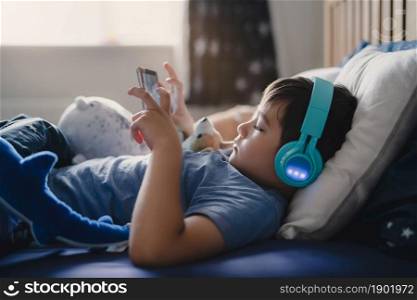 Cute young boy wear wireless headphone listening music while lying in bed, Happy kid playing game on tablet,Positive child in blue pajamas relaxing in bed room in morning before go to schoo