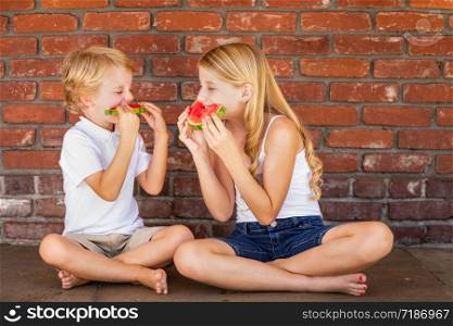 Cute Young Cuacasian Boy and Girl Eating Watermelon Against Brick Wall.