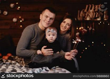 cute young family dad, mom and baby girl with sparklers on bed with Christmas background behind. cute young family dad, mom and baby girl with sparklers on bed with Christmas background behind.