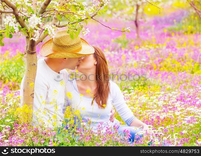 Cute young family sitting down on pink floral meadow in the garden and kissing, woman and man enjoying each other outdoors, romance concept
