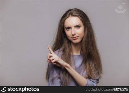 Cute young female model with brunette long hair, points at blank copy space of grey wall, advertizes something with serious expression, attracts your attention. Advertisment and people concept