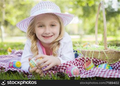 Cute Young Girl on Picnic Blanket Wearing Hat Enjoys Her Easter Eggs Outside in the Park.