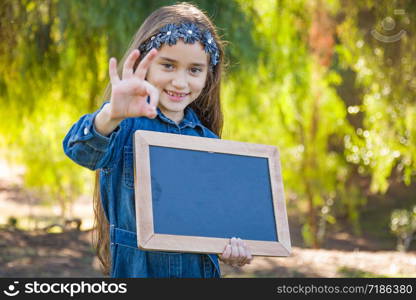 Cute Young Mixed Race Girl With Okay Sign Holding Blank Blackboard Outdoors.