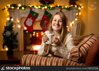 Cute young woman sitting at fireplace with cup of tea at Christmas