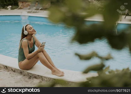 Cute young woman sitting by the swimming pool and listen music from mobile phone with headphones in the house backyard
