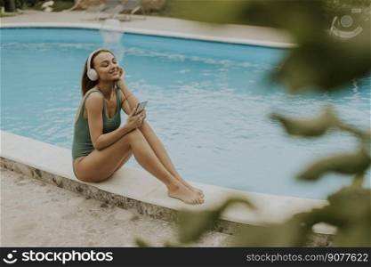 Cute young woman sitting by the swimming pool and listen music from mobile phone with headphones in the house backyard