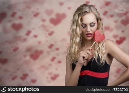 cute young woman with long wavy blonde hair and sexy dress taking heart shaped lollipop in the hand