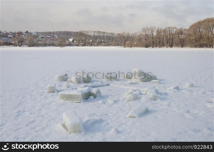 Cutted out cubes of ice on frozen lake near the hole. Village in the distance.
