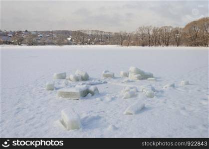 Cutted out cubes of ice on frozen lake near the hole. Village in the distance.
