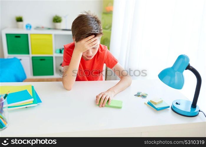 cyberbullying, technology and people concept - tired or suffering from headache or bullying student boy with smartphone sitting at desk at home. tired student boy with smartphone at home