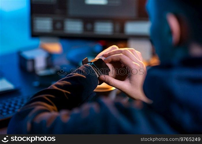 cybercrime, hacking and technology concept - close up of male hacker in dark room with smart watch and computer carries out cyber attack in dark room. hacker with smart watch and computers in dark room