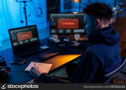 cybercrime, hacking and technology concept - male hacker in dark room writing to notebook and using computer virus program for cyber attack. hacker using computer virus for cyber attack