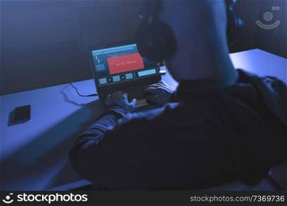 cybercrime, hacking and technology concept - male hacker in headphones with access denied message on laptop computer screen using virus program for cyber attack in dark room. hacker with access denied message on laptop