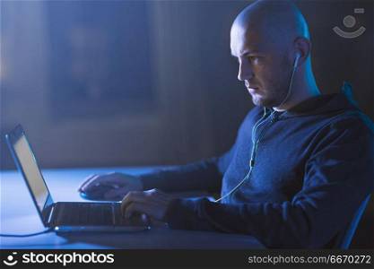 cybercrime, hacking and technology concept - male hacker with earphones using laptop computer for cyber attack in dark room. hacker in earphones typing on laptop in dark room. hacker in earphones typing on laptop in dark room