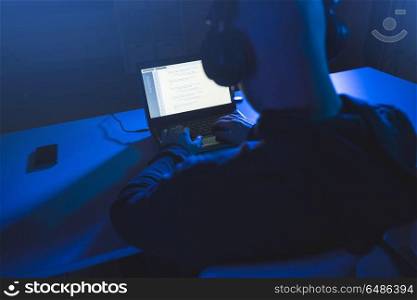 cybercrime, hacking and technology concept - male hacker with headphones and coding on laptop computer screen wiretapping or using computer virus program for cyber attack in dark room. hacker with coding on laptop computer in dark room. hacker with coding on laptop computer in dark room
