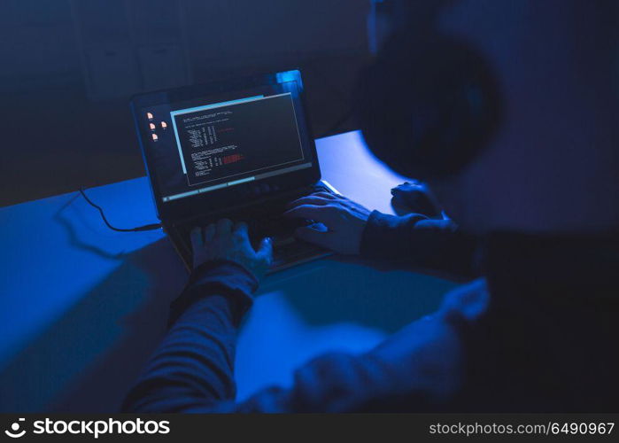 cybercrime, hacking and technology concept - male hacker with headphones and coding on laptop computer screen wiretapping or using virus program for cyber attack in dark room. hacker with coding on laptop computer in dark room. hacker with coding on laptop computer in dark room