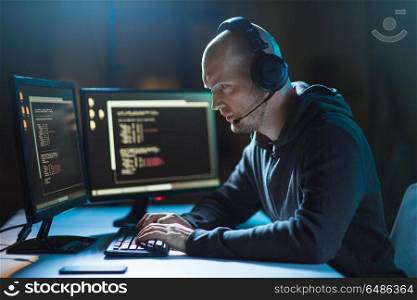 cybercrime, hacking and technology concept - male hacker with headset and coding on computer screen wiretapping or using computer virus program for cyber attack in dark room. hacker with computer and headset in dark room. hacker with computer and headset in dark room